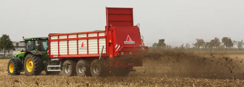 Czerwono-biały rozrzutnik obornika Annaburger na podwoziu trzyosiowym, ciągnięty przez traktor John Deere podczas pracy na ściernisku.
